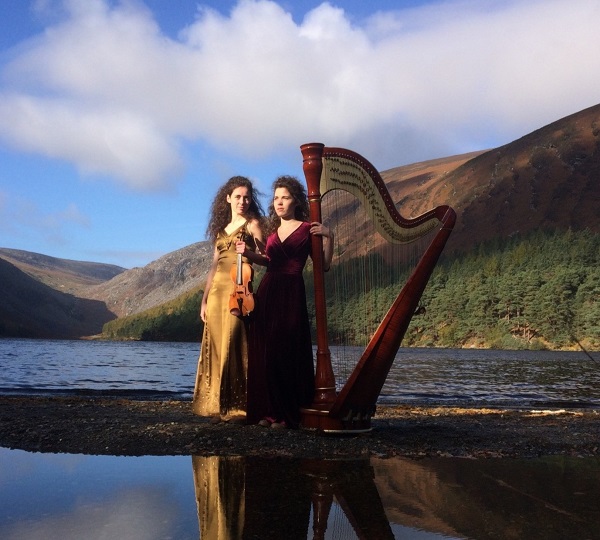 Les 2 membres du groupe et une harpe au bord d'une étendue d'eau avec des montagnes en fond.